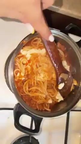The video shows a top-down view of a small stainless steel pot filled with spaghetti, red sauce, and globs of white cheese on a white stovetop. A hand with white and yellow nail polish uses a brown wooden spoon to stir the ingredients together. Steam is visible rising from the pot, indicating that the food is hot.