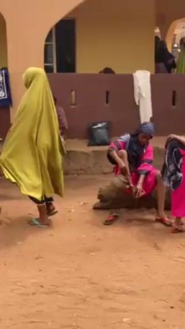 The video shows a gathering of people in what appears to be a village or rural area with predominantly reddish-brown dirt ground. Many individuals are wearing traditional or modest clothing including hijabs. One woman in a bright pink outfit and hijab is sitting on a stone structure or ledge, and appears to be cleaning or washing her foot with water from a jug. Another woman in yellow robes walks past her. Several men are seen walking through the scene; one is wearing a blue and yellow shirt, dark pants, and a small blue cap. He gestures towards the distance, where several wheeled carts with food or goods are visible. Several people are standing around watching the woman with her foot. The environment appears to be a public space, possibly near buildings or homes.