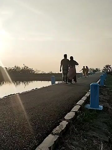 The video depicts an outdoor scene in Jambeyan, Indonesia, likely during either sunrise or sunset due to the low angle of the sun and the strong backlighting. Several people are seen walking along a paved or gravel path that runs parallel to a calm body of water, possibly a canal or reservoir. In the foreground, two figures, silhouetted against the bright sky, walk away from the camera. Further along the path, additional silhouetted figures are visible, also walking. The path is bordered on the right by small, blue and white painted bollards or markers. Sparse vegetation and trees can be seen along the water's edge and in the distant background. The sky is clear, and a sun flare is visible on the left side of the frame, indicating direct sunlight. No visible text is present in the video.