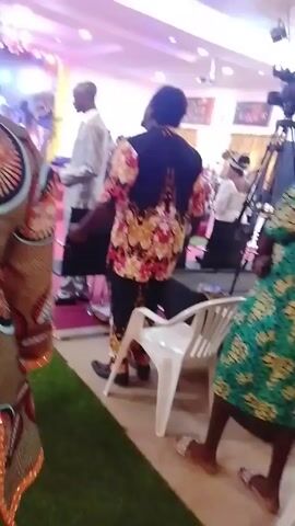 The video appears to be shot at an event, possibly a church service or ceremony, with a focus on individuals in the foreground and background. In the foreground, several people are visible, including women wearing colorful patterned dresses and men in more traditional attire, such as a white outfit. One woman in a green and yellow dress is seen near a white plastic chair. The camera briefly moves to capture other people. The environment is a large indoor space with bright lighting, and there are other people and objects in the background, including a video camera on a tripod. The event seems lively.