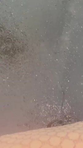 The video shows a person walking on a wet surface, likely asphalt, during what appears to be a rain shower. The camera is positioned at a low angle, focusing on the person's feet and legs as they walk. The person is wearing dark, strappy sandals and light-colored pants with a repeating pattern of interlocking Gs. As the person walks, their sandals splash water, creating ripples on the surface. The ground is dark and appears to be littered with small debris. In the later frames, yellow markings, possibly a crosswalk or lane divider, become visible on the wet surface. The overall impression is of someone walking through puddles in the rain.