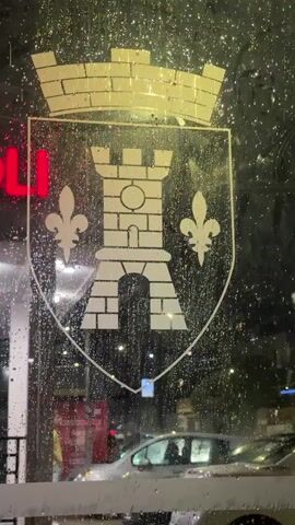 The video shows a view through a window covered in water droplets, suggesting it is raining. A crest with a castle and fleur-de-lis is visible on the glass. Outside, a neon sign with red letters spelling "VOLI" is lit. Cars are parked on the street, and streetlights illuminate the scene. The overall atmosphere is dim and wet.