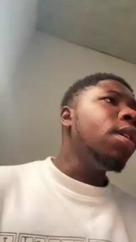 A close-up shot features a young Black man looking upward and then directly at the camera. He wears a white sweatshirt with printed text and images on it. His hair is short and dark, and he has a short beard. The background is a light-colored wall. Music can be heard in the background.