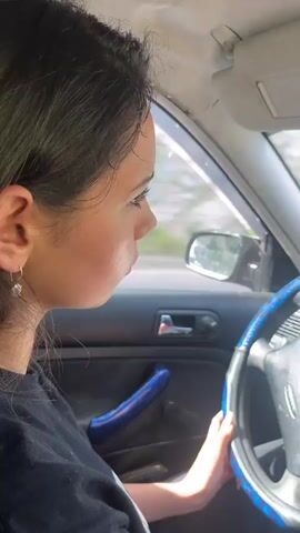 The video is a first-person perspective from inside a car. A woman with dark hair is driving, her hands are on the steering wheel. The car's interior is visible, including the dashboard with a can of Coca-Cola in a cup holder and a rearview mirror with a hanging air freshener. The road ahead is visible through the windshield, showing a winding road bordered by rocky cliffs and lush green trees. The sun is shining, and the sky is blue with some clouds. The car appears to be in motion.