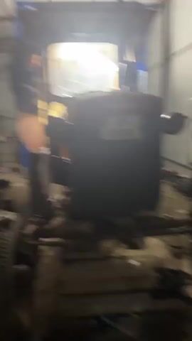 A young man is working on a tractor inside a garage. The tractor is a dark color with a yellow cabin. The man is standing on the side of the tractor, near the tire. He is wearing a dark jacket and pants. He turns towards the camera and sticks out his tongue and points at the camera. The garage is cluttered with tools and equipment. The light is bright inside. Another person is working on the side behind the tractor.