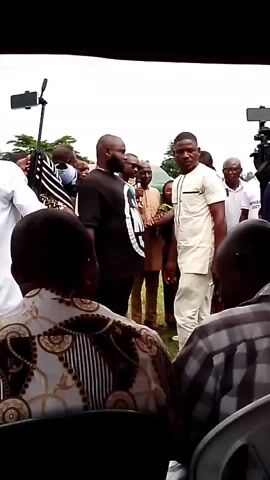 A group of people are gathered outdoors, with several individuals facing the camera. The central focus is a man in a light-colored traditional outfit, standing and speaking. To his left, another man in a black outfit is also facing him, and they appear to be interacting. In the background, more people are visible, some holding up phones, suggesting recording or photography. There are also cameras and possibly media equipment present. The environment appears to be an open area, possibly a gathering or event, with trees and greenery visible in the distance. The sky is overcast.