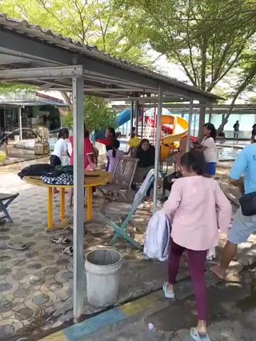 The video shows a lively scene at what appears to be a public swimming pool. A group of children and some adults are gathered under a covered structure with tables and chairs. Some of the children are changing clothes or organizing their belongings, while others are heading towards the pool area. The pool itself is visible in the background, with several people swimming or playing in the water. The overall atmosphere seems relaxed and recreational. The ground is covered with patterned tiles, and trees are visible outside the covered area, suggesting an outdoor setting.