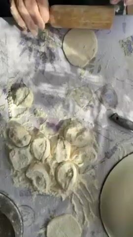 A person is preparing dough on a lavender-patterned tablecloth covered in flour. They use a wooden rolling pin to flatten a small, round piece of dough. There are several other similar, unrolled pieces of dough nearby, also covered in flour. A knife and a white bowl are visible on the tablecloth to the side. The scene is well-lit, with natural sunlight creating shadows across the surface. The person's hands and part of their torso are visible.