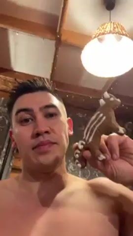 A shirtless man holding a gingerbread cookie shaped like a raccoon takes a bite. The man is fair-skinned with short dark hair. He is indoors, with a ceiling overhead that appears to have wooden beams, a light fixture, and decorative lights. The background includes curtains or drapes. The man looks directly at the camera.