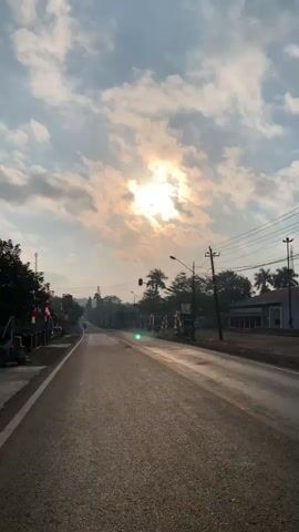 The video shows a forward-facing perspective of a paved road with a white line marking the left lane. The road appears to be a rural or suburban area, with trees and some buildings visible on both sides. The sun is visible in the upper portion of the frame, partially obscured by clouds, casting a bright light and creating some lens flare. There are power lines running overhead, supported by utility poles. Red and white striped flags are visible along the side of the road. A few vehicles, including what appears to be a truck, are parked or moving in the distance. The overall atmosphere suggests an outdoor scene during daytime, possibly early morning or late afternoon given the lighting.