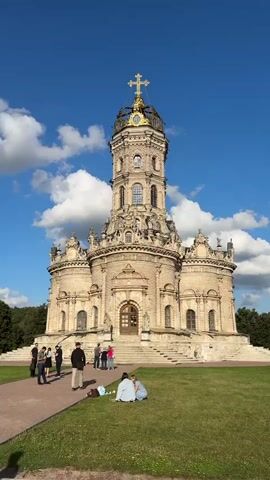 The video begins with a wide shot of a large, ornate church with a tall, multi-tiered bell tower topped with a golden cross. The church is made of light-colored stone and features intricate carvings and arched windows. The sky is blue with scattered white clouds. In the foreground, several people are gathered on a grassy area in front of the church. Some are standing and talking, while others are sitting on the grass. The camera slowly zooms in on the church, focusing on the detailed architecture of the bell tower. The video then pans across the church's facade, revealing more of its decorative elements. Finally, the video zooms out slightly, showing the church and the surrounding landscape with a few more people visible in the distance. The overall scene conveys a sense of grandeur and tranquility.
