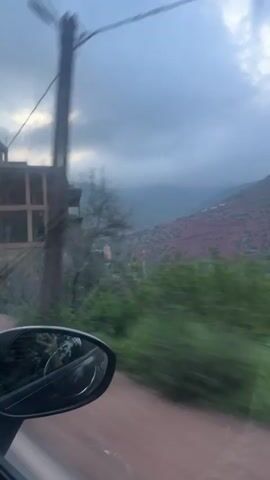 The video captures a view from inside a moving vehicle, primarily looking out the passenger side window and occasionally through the windshield. The scene depicts a journey through a mountainous region, consistent with the provided location of Asni, Morocco.

Initially, the view shows an unfinished, multi-story concrete building with exposed rebar on the left side of the road, set against a backdrop of distant mountains under a cloudy, overcast sky. Lush green trees, some blurred due to motion, are visible between the car and the building. A power line stretches across the top of the frame. The car's side mirror is prominent in the foreground, reflecting the sky and passing scenery.

As the vehicle continues, the landscape shifts to reveal red-brown brick buildings lining the narrow road, suggesting passage through a village or town. One shot shows a close-up of a brick wall from the car's interior. Further down the road, more brick structures are visible, and a white car can be partially seen. The final shot from the driver's perspective shows the winding road ahead, flanked by similar red-brown buildings and distant mountains, with lights visible further along the path, possibly indicating an illuminated area or a tunnel entrance.

The overall impression is that of a road trip through a developing area in a mountainous, often overcast, landscape. There is no visible text in the video.