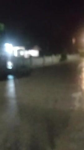 The video captures a nighttime scene on a wet, asphalt road. A street light illuminates the road and the surrounding area, reflecting off the wet surface. There are faint outlines of buildings and a fence on the left side of the road. The overall atmosphere is dark and the visuals are blurry, suggesting low light conditions and possibly motion.