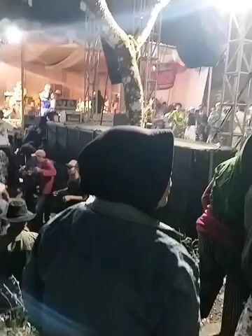 A concert is taking place at night. A band is on stage performing for a crowd of people. Many members of the audience are standing and some are dancing. There are trees and lights in the background. The camera is handheld and shaky, suggesting it was filmed by someone in the audience. The lighting is dim and there are some purple and pink lights on stage. The overall mood is festive and energetic.