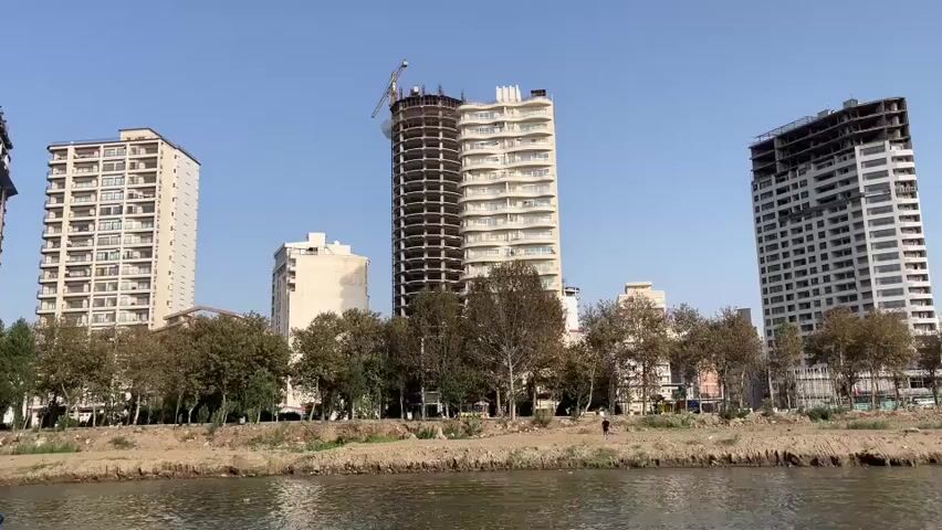 The video shows a wide shot of a river with a cityscape in the background. Three tall buildings are visible, one of which is under construction with a crane at the top. A line of trees separates the river from the buildings. Two boats are on the water. The boat in the foreground is white with a white canopy and appears to be a tour boat with passengers. A smaller, colorful boat with people on board is moving quickly to the right in the middle ground. The sky is clear and blue.