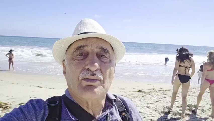 A man wearing a white fedora and a backpack takes a selfie on a sandy beach. The ocean and waves are visible in the background. Two women in bikinis are walking away from the camera towards the water, holding hands. Another woman in a bikini is standing closer to the water on the left side of the frame. Several other people are visible in the distance. The sky is clear and blue.