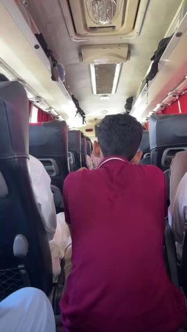 The video displays the interior of a passenger bus, viewed from a seat towards the front. The primary subject is the back of a man with dark hair, wearing a maroon polo shirt with a white collar stripe, seated in front of the camera. To his left, another passenger in white clothing is partially visible.

The bus features black-upholstered seats arranged in rows, red curtains along the windows, and white overhead panels. Integrated into the ceiling are long light fixtures, air vents, and overhead luggage racks that hold various items, possibly bags or clothing. Black straps hang from these racks.

At the very front of the bus, above the driver's area, a small display screen is visible. It shows the time as "10:07" and a partial number "0315-7xxx", which might be a contact number or route identifier. Several other passengers are seated down the aisle.

The bus appears to be in motion, suggested by the general atmosphere and sounds. The bright interior lighting and the time displayed indicate it is daytime. The setting is consistent with public transportation in Chak 582 GB, Pakistan. Emotions of the passengers are not discernible from this perspective.