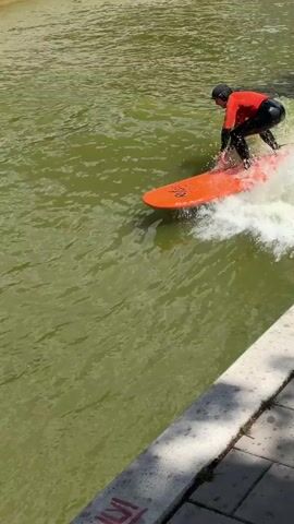 A person wearing a red long-sleeved shirt and black full-body wetsuit is surfing on an orange surfboard in a river with greenish-brown water. The person is wearing a black helmet and is standing with their back to the camera, facing the direction of the wave. White water is splashing around the surfboard. In the background, there is a concrete embankment with a blurred crowd of people and structures visible. The water appears somewhat choppy, with a consistent wave being ridden.