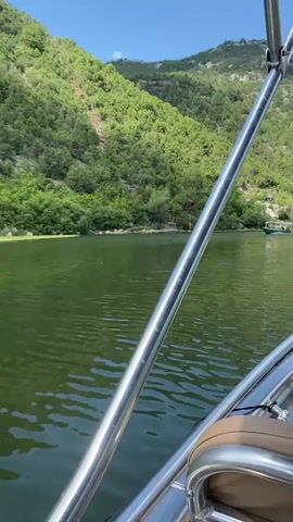 The video is a first-person view from a boat moving across a calm, green body of water. On the left side of the screen, a lush, green mountainside rises steeply, covered in trees. A dirt path is visible snaking up the mountainside in the distance. On the right side of the frame, a green boat with a white canopy is moving in the same direction as the camera's boat. The boat has people on board. The water is calm with small ripples. The sky is clear and blue.