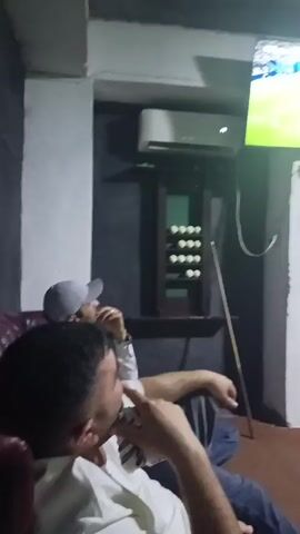 The video shows two people watching a soccer match on a television. A man in a white shirt and blue jeans is seated in the foreground, his face turned towards the television. To his left, another person wearing a grey cap is partially visible. The television screen displays a soccer field with players. In the background, an air conditioning unit is mounted on the wall above a dark wooden cabinet filled with billiard balls. A billiard cue leans against the wall near the cabinet. The room appears to be dimly lit.