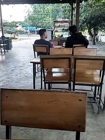 The scene shows a semi-outdoor restaurant setting with two people sitting at a table. The restaurant has a concrete floor and a metal roof, providing shade. There are several empty wooden tables and chairs in the foreground. The two people are sitting at a table further away, engaged in a conversation. One person is wearing a dark blue shirt and the other is wearing a black jacket. Beyond them, a food cart is visible in the background, along with some trees and buildings. The overall atmosphere is quiet and relaxed.