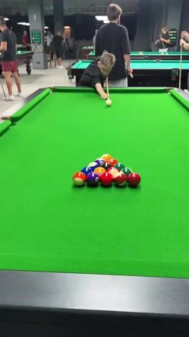 A group of people are in a pool hall, playing billiards. In the foreground, a young boy in a black t-shirt is lining up a shot on a pool table. A rack of pool balls is set up in the center of the table. The boy is holding a pool cue and appears to be about to strike the cue ball. Other people are visible in the background, some playing pool at other tables and others watching. The pool hall appears to be well-lit with a green felt pool table.