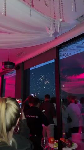 This media file captures a festive scene from what appears to be a rooftop or elevated venue, likely in Lisi, Georgia. The time of day is dusk or early evening, as indicated by the dark sky punctuated by bright fireworks.

The primary subjects are a group of people, silhouetted against the window, observing the fireworks display. Many are holding up their phones to record or photograph the event, suggesting excitement and a desire to capture the moment. The mood seems celebratory and engaging. The setting is indoors, with a white draped ceiling and pink ambient lighting casting a warm glow on the scene. Large windows offer a panoramic view of the night sky where colorful fireworks are exploding.

Notable details include the vibrant fireworks, with bursts of yellow, red, and white light illuminating the sky. The reflection of the fireworks and the interior lighting can be seen on the large windows. There is a decorative element hanging from the ceiling, possibly a pearl-like garland with a translucent bird ornament. The people are casually dressed, further contributing to the relaxed, celebratory atmosphere.

The activity taking place is the viewing of a fireworks display. One person in the foreground wears a black jacket with what appears to be text in Georgian script on the back. While not fully legible, the text is red and stands out against the dark fabric. The overall impression is one of shared enjoyment of a special event.