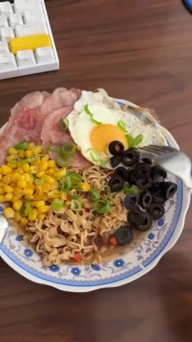 A plate of food sits on a wooden table. The food consists of noodles, corn, ham, a fried egg, black olives, and chopped green onions. A fork is being used to eat the food. A keyboard is visible in the upper left corner of the image.