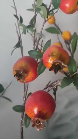 This is a close-up, static shot of several small, artificial pomegranates and a few small yellow fruits, possibly kumquats, attached to branches with green leaves. The pomegranates are red and orange, with darker speckles and a visible calyx at the bottom. The yellow fruits are round and bright. The branches are thin and brown, with a few scattered leaves. The background is a plain, light-colored wall. The overall impression is one of artificial decorative fruit. There is no indication of people, weather, time of day, or emotions. There is no visible text in the image. The location context of Batken City, Kyrgyzstan is not directly inferable from the visual content of the image itself.