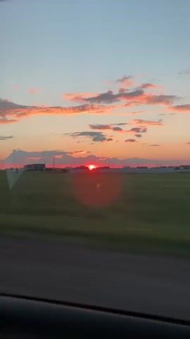 A time lapse video captures a sunset viewed from inside a vehicle. The sun, a bright red orb, dips below the horizon, casting a vibrant orange and pink glow on the scattered clouds. The landscape consists of a vast, flat green field with a few houses visible in the distance. The road in the foreground is blurred due to the motion of the vehicle. The sky transitions from a pale blue at the top to shades of orange and pink as it nears the horizon.