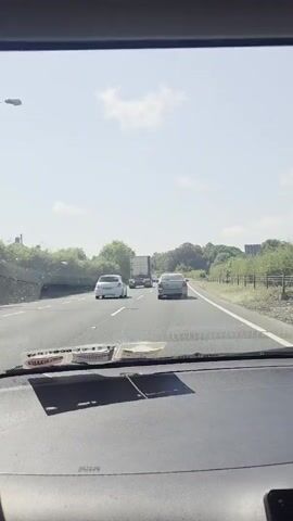The video shows a view from inside a car driving on a highway. The car is on the right lane, and other vehicles are visible on the highway ahead. A white car is in the left lane, and a truck is in front of it. Further ahead, a silver car is in the right lane. The sky is clear with some clouds, and there are trees and bushes on the sides of the highway. A speed limit sign indicating 100 is visible on the right side of the road. The dashboard of the car is visible in the foreground.