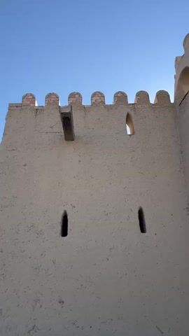 The video shows a low-angle shot of a section of an old, tan, mud-brick wall against a clear blue sky. The wall has crenellated battlements along the top and a series of narrow, vertical slits that appear to be windows or arrow slits. A wooden beam protrudes from the wall near the top. As the camera pans to the right, the wall continues, revealing more of its length and showing small wooden beams projecting from it at intervals. The wall is bordered by vegetation and a metal fence in the foreground. In the distance, a part of a more modern structure with beige walls and a tan roof is visible. The overall impression is that of an ancient fortification.