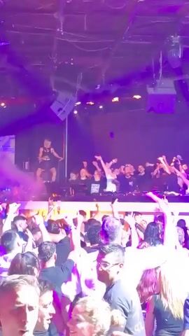 A large crowd of people, mostly men, are dancing at a nightclub. There is a DJ performing on an elevated stage with flashing lights. The club has a dark interior, and the lighting consists of multiple spotlights of varying colors. The crowd is packed together, and people are raising their hands in the air. Fog or smoke is visible, which adds to the atmosphere of the nightclub.