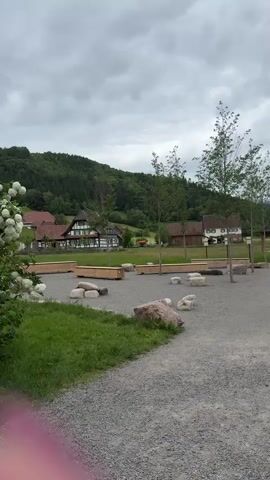 Two women are sitting on a bench alongside a gravel path in a scenic rural area. The path leads towards a gazebo and a fenced enclosure with buildings beyond. The sky is overcast, and the surrounding landscape features rolling hills covered in trees. A sign beside the bench invites visitors to a "Winzerstube" (wine tavern) open daily from 11:00. A pile of firewood is also located near the bench.
