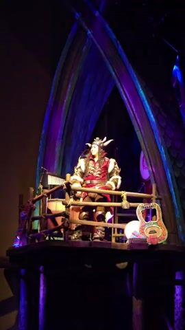 This image captures a performer dressed as a Viking on a stage set, likely part of a theatrical production or theme park attraction. The Viking is wearing a red tunic over armor, a horned helmet, and fur accents, exuding a fierce yet theatrical presence. They are positioned on a wooden platform, which appears to be elevated and surrounded by rustic railings.

The stage is adorned with various props, including what looks like a xylophone or metallophone, drums, and a uniquely designed stringed instrument with colorful carvings. The backdrop features a large, arched structure with what resembles scales, illuminated by vibrant purple and blue stage lighting, creating a dramatic and fantastical atmosphere. This lighting suggests the performance is taking place indoors, possibly in the evening or a darkened venue, and the overall mood is theatrical and immersive.

The setting evokes a sense of ancient or mythical times, fitting for a Viking theme. The performer's posture and the context of the stage suggest they are either singing, speaking, or engaging in some form of performance. There is no visible text in the image. The location context of Tongzhou District, China, indicates this might be a cultural performance or entertainment venue within that region.