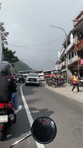 The video shows a view from a motorcycle in traffic on a road lined with buildings. The road is paved and has a white line dividing the lanes. A white SUV is visible in the foreground, followed by other cars and motorcycles. On the right side of the road, there are several buildings with shops or restaurants, and many motorcycles are parked along the sidewalk. The sky is overcast, and there are trees and hills in the background. The overall scene depicts a busy street in a town or city, possibly a tourist area, with a lot of vehicle traffic.