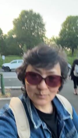 The video shows a woman with short, dark curly hair and sunglasses, wearing a denim jacket and a backpack. She is standing on a paved path with a road and grass on either side. There are trees in the background and cars parked on the road. She is looking at the camera and smiling slightly. The camera is held by the woman, creating a selfie perspective. A person walks by in the background.
