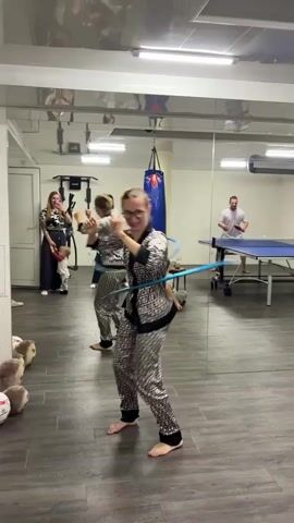 The video shows a group of people in a room with a large mirror on one wall. In the foreground, a young woman with blonde hair in a bun, wearing a patterned tracksuit, is hula hooping. She is barefoot and focused on keeping the hoop spinning. Behind her, another young woman in a similar tracksuit is also hula hooping, though slightly out of focus. To the left, a woman holding a phone is watching, with a small child standing next to her. Further back, a man in a white t-shirt is standing near a ping pong table, holding a paddle. A punching bag hangs from the ceiling in the background. The room appears to be a recreational space, possibly a gym or a party venue, with workout equipment visible on the left side of the mirror. The floor is a dark, reflective surface.