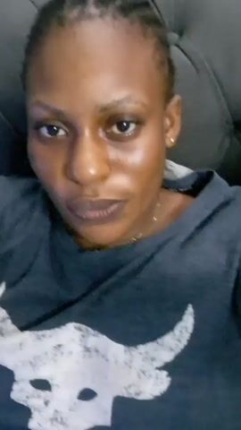 A woman with dark hair pulled back in braids and wearing a gray t-shirt with a white design is lying on a gray tufted couch. She is looking directly at the camera. A gold chain is visible around her neck, and she has gold stud earrings.  A hand reaches into the frame momentarily, then withdraws. In the background, a water cooler, an electric heater, and a filled blue water jug are visible.  A child's voice is briefly heard.