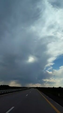 The video shows a view from a car driving on a highway. The sky is filled with dark, dramatic clouds, with a small patch of blue sky visible on the right side. There appears to be some light rain or mist falling in the distance on the left side of the frame. The road ahead is empty except for a few distant vehicles. The scene is dimly lit, suggesting it might be late afternoon or early evening, or that the clouds are obscuring the sun. The environment appears to be rural, with flat, dark terrain on either side of the road. The video is short, lasting only a few seconds, and the camera is stationary, implying it was recorded from a fixed position within the vehicle.