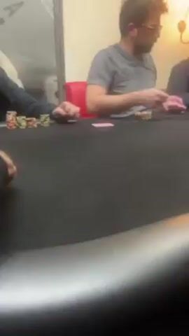 The video shows a scene from a poker game taking place at a black felt-covered poker table. Three men are visible seated around the table, each holding cards. The closest player is wearing a gray T-shirt and glasses, while another is wearing a dark navy blue shirt. Chips are stacked in front of the players, and a pack of cigarettes sits on the table. The environment appears to be indoors with lighting from an unseen source in the upper right-hand corner. A red chair is situated behind the player with the gray T-shirt. In the background, there's a wall with an Ace of Spades decoration.