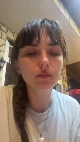 The video begins with a close-up shot of a young woman with dark hair and bangs, her eyes closed, holding a small, tulip-shaped glass of dark red liquid. She is wearing a white t-shirt and a necklace with a white heart-shaped pendant. Her left braid is visible over her shoulder. The background appears to be an outdoor seating area with stone walls and a light fixture.

The woman brings the glass to her lips and takes a sip. Her eyes flutter open as she drinks, and she then closes them again. After finishing the drink, she lowers the glass and a faint smile appears on her lips. The camera remains focused on her face throughout the short clip.