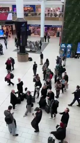 The video shows a high-angle view of an airport terminal lobby. Numerous people are scattered throughout the area, some standing in groups, others walking with luggage. Many are looking at their phones or checking flight information screens, which display flight details and departure times. Shops and food vendors can be seen on a second level, visible above the crowd. The floor is covered with large, light-colored tiles. There are several support pillars, one of which has flight information screens wrapped around it.