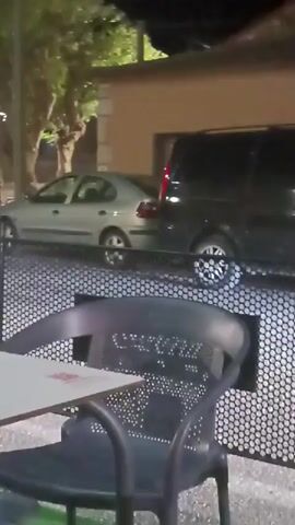The video shows an outdoor setting at night, possibly a cafe or restaurant, with tables and chairs visible. There are cars in the background, one of which appears to be driving over some concrete blocks. Several people are seated at tables, some facing the camera and others with their backs to it. The lighting is dim, with streetlights and interior lights illuminating the scene. The overall atmosphere appears to be casual and social, with people engaged in conversation.