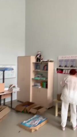 This media file shows an indoor scene, likely a classroom or office, in Megion, Russia. The room is brightly lit, suggesting daytime.  The walls are a pale blue, and the ceiling is a white drop ceiling with fluorescent lighting fixtures.

In the foreground, a person wearing a white shirt and white pants is standing and appears to be interacting with a cardboard box. To the left, there is a light-colored cabinet with shelves filled with books and other items. On top of the cabinet, a small doll or puppet dressed in a suit and hat is visible. A poster with Russian text, including "ПАРАД РЕЗУЛЬТАТОВ," is mounted on the wall. Other posters with what appear to be certificates or awards are also displayed. Several cardboard boxes are on the floor. In the background, vertical blinds cover a large window.

The activity seems to be organizing or packing items, as evidenced by the boxes and the person's actions. The overall impression is one of a functional, possibly educational or administrative space.