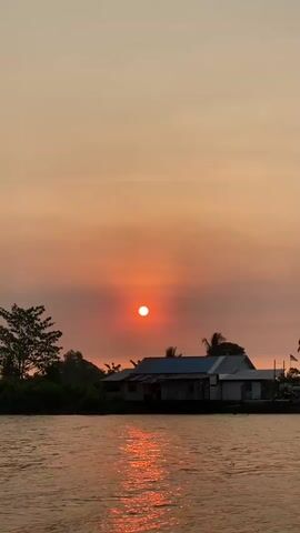 The video shows a sunset scene over a body of water. In the foreground, there is water reflecting the orange hues of the sunset. In the middle ground, there are some buildings and trees on the bank. In the background, a large yellow suspension bridge spans the water. The sky is a gradient of orange and yellow, with the sun appearing as a bright orange orb partially obscured by haze. The overall impression is a peaceful, natural landscape.