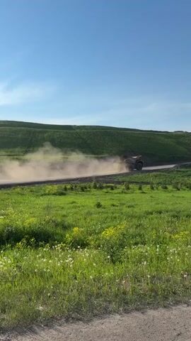 A large, tan dump truck, heavily laden with dirt and rocks, moves from left to right on a dirt road, kicking up a large cloud of dust behind it. The truck is centered in the frame. The road is bordered by a small, dark dirt bank on the right, and then by a large field of green grass with small yellow and white flowers on both sides. In the distance, a large, green, grassy hill stretches across the background under a clear, bright blue sky. The sunlight appears to be coming from the left, casting a slight shadow under the truck and illuminating the dust cloud.