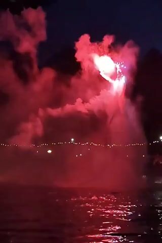 A nighttime scene shows a body of water illuminated by a bright pink flare. The flare is positioned above and slightly to the right of the center of the frame, emitting a dense cloud of pink smoke that billows outwards. The water reflects the pink light, creating a shimmering effect. In the background, a line of blurry lights suggests a distant shoreline or structures. To the left of the frame, a jet ski is visible on the water, also illuminated by the pink light, with a faint green light on its rear. The rider is not clearly visible. The overall atmosphere is dramatic and festive.