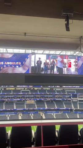 This media file captures a wide shot of a stadium interior, likely during a daytime event, as indicated by the bright lighting. The primary focus is on a large, high-definition screen displaying a live feed or pre-recorded content.

The screen shows a group of individuals performing, appearing to be a musical ensemble, possibly a band or orchestra. Several members are playing instruments such as trumpets and drums. Behind them, the backdrop features bold text that reads "LOS ANGELES" in large, stylized font, accompanied by decorative elements resembling stars and wavy lines, suggesting a celebratory or promotional theme related to the city. The screen also displays "FIFA WORLD CUP 2026" and "LOS ANGELES" on other parts of the frame.

Below the large screen, several tiers of stadium seating are visible, mostly empty. The seats are dark blue with some red accents visible in the foreground. The overall atmosphere appears to be one of anticipation or a break in the main event, with a diverse audience present. A few individuals are seated in the foreground, looking towards the screen. The stadium setting and the "FIFA WORLD CUP 2026" branding strongly indicate the location is Inglewood, United States of America, likely SoFi Stadium, which is a prominent venue for major sporting events and concerts. The clear, bright lighting suggests it's daytime.
