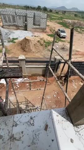 The video presents an elevated view of an active construction site in Angwan Sayawa, Nigeria, on an overcast day.

In the immediate foreground, a white, marble-patterned tiled floor or balcony is visible, strewn with dirt and construction debris. A green bucket containing tools and a long metal straight edge tool rest on this surface. Looking down from this vantage point, a complex scaffolding structure made of wooden poles and metal supports extends downwards and outwards. A long wooden plank, heavily coated with fresh concrete or mortar, is positioned on the scaffolding. A metal ladder leans against the structure.

In the mid-ground, a dark-skinned male worker, dressed in dark clothing, is seen sitting or crouching near the base of the scaffolding, possibly resting or performing a low-level task. The ground around him is reddish-brown earth.

Further into the scene, an unfinished building constructed from concrete blocks, lacking a roof and windows, stands prominently. Nearby, piles of sand and other construction rubble are visible, alongside a parked white car. A concrete fence with decorative pillars runs across the landscape, and a tall solar street light is positioned behind it.

The background shows a sprawling development area with numerous other partially constructed buildings, indicating ongoing large-scale construction. Distant hills or mountains frame the horizon under a hazy, bright sky. The overall scene depicts a raw, developing construction zone filled with materials and infrastructure typical of building projects.